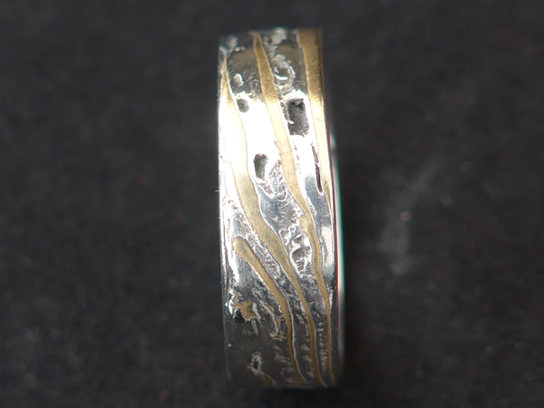 Deze stoere ring is gemaakt van Mokume Gane. Dit is een bewerkelijke Japanse smeedtechniek waarbij laagjes verschillende metalen, in dit geval zilver en koper, met elkaar versmelten onder hoge temperaturen. In het Japans betekent mokume houtnerf en gane betekent metaal. 